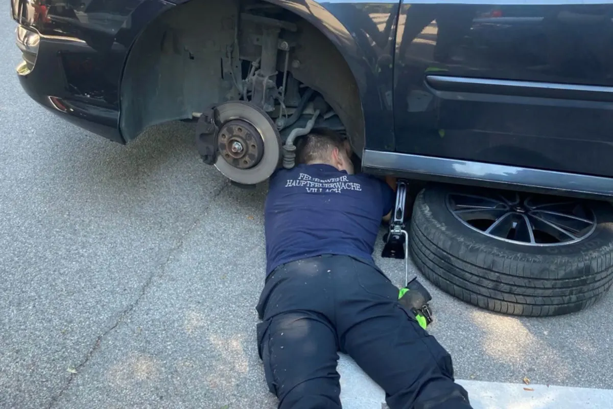 Eine Person in einem Feuerwehrshirt arbeitet unter einem angehobenen Auto. Das Fahrzeug steht auf einem Wagenheber am Straßenrand.