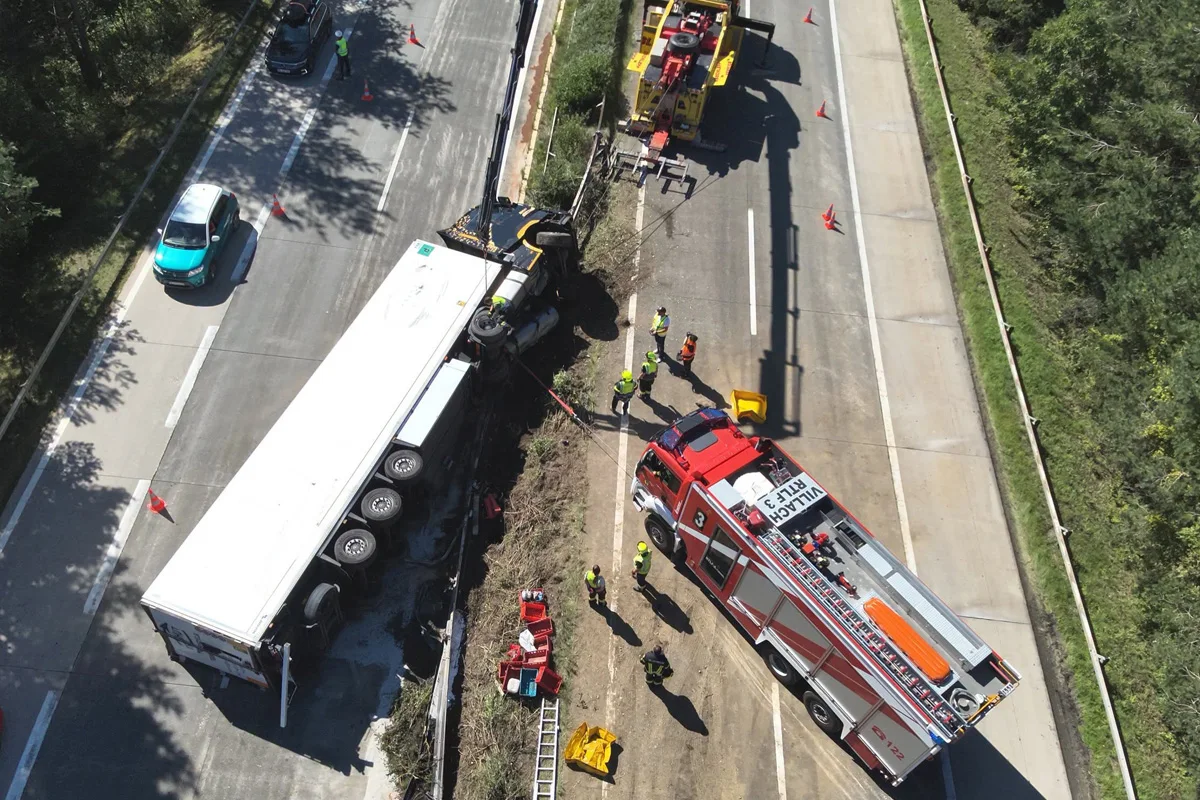 Ein großer Lkw liegt auf der Seite einer Straße, während mehrere Einsatzkräfte und ein Feuerwehrauto vor Ort sind, um die Bergung zu unterstützen.