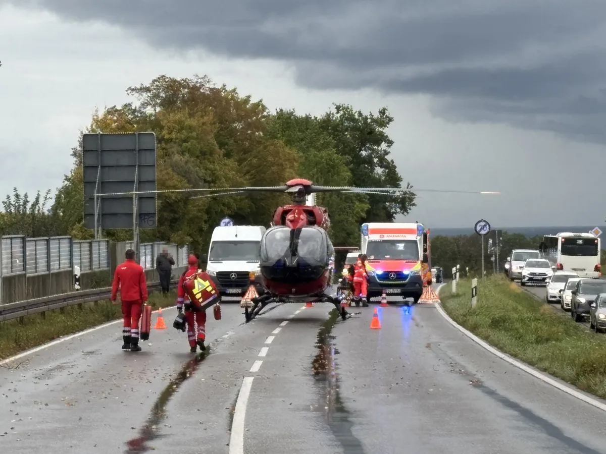 Ein Rettungshubschrauber steht auf einer nassen Landstraße, umgeben von Rettungskräften in roten Anzügen und einem Rettungswagen.