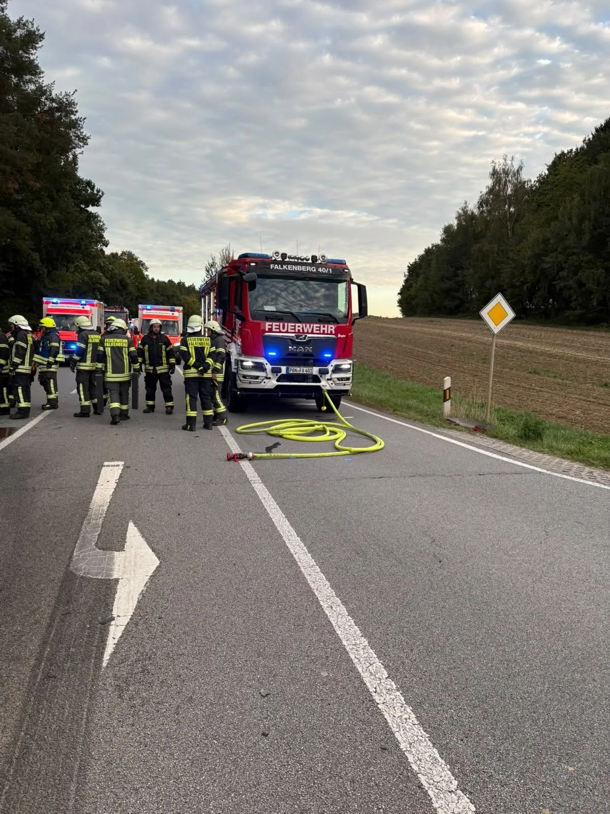 Ein rotes Feuerwehrfahrzeug mit der Aufschrift 'Feuerwehr' steht auf einer Landstraße. Mehrere Feuerwehrleute in Schutzausrüstung sind um das Fahrzeug herum versammelt. Ein gelber Schlauch ist auf der Straße ausgerollt.