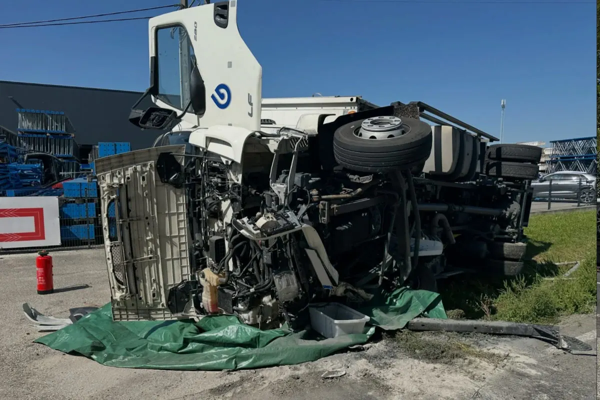 Ein großer weißer Lastwagen liegt auf der Seite, seine Mechanikteile sind freigelegt. Im Hintergrund befinden sich Metallgerüste.
