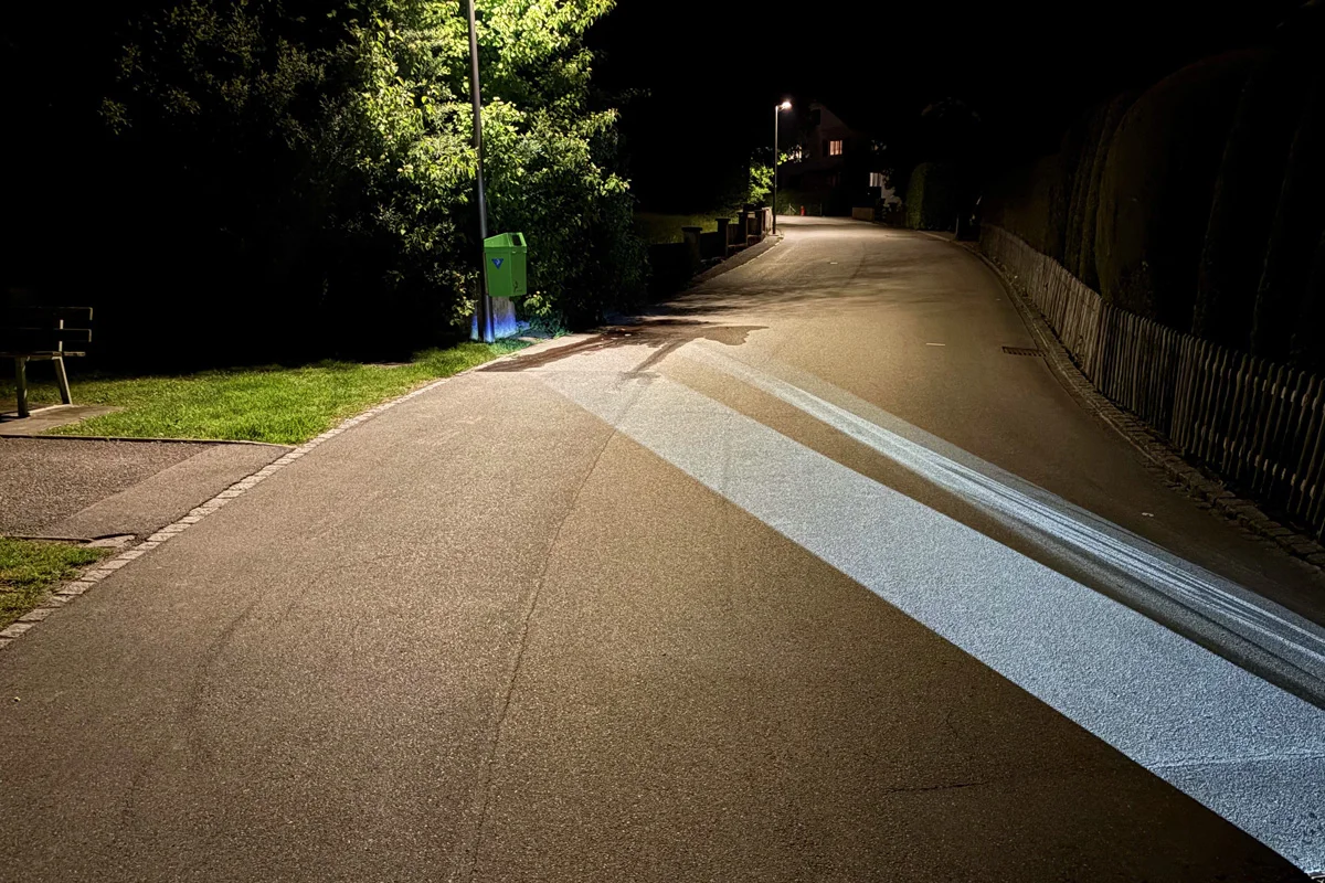 Eine Straße bei Nacht mit zwei intensiven Lichtstrahlen, die diagonal die Fahrbahn kreuzen. Bäume und Mülleimer sind auf dem Gehweg sichtbar.