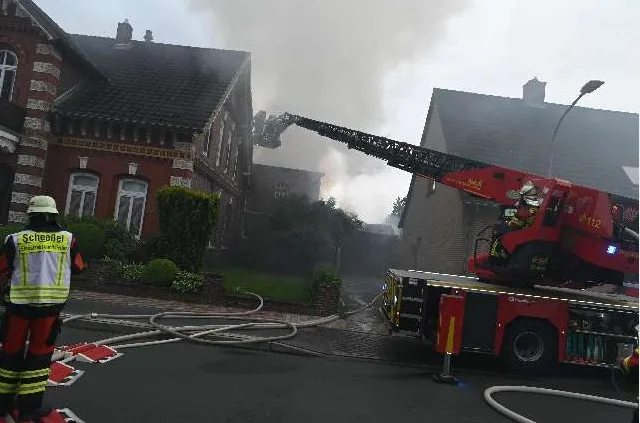 Ein Feuerwehrfahrzeug mit Drehleiter steht vor einem brennenden Haus, aus dem dichter Rauch aufsteigt. Ein Feuerwehrmann ist im Einsatz.