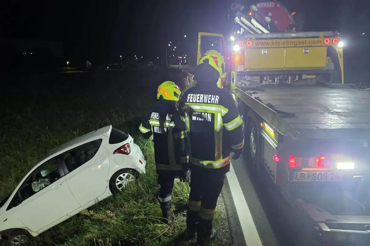 Zwei Feuerwehrleute stehen neben einem weißen Auto, das von der Straße abkam und im Graben liegt. Ein Abschleppfahrzeug ist ebenfalls vor Ort.