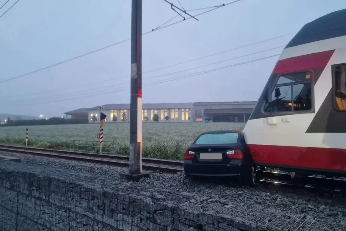 Ein Personenzug steht frontal vor einem schwarzen Auto auf den Gleisen. Es herrscht Dämmerung, und ein Gebäude ist im Hintergrund sichtbar.