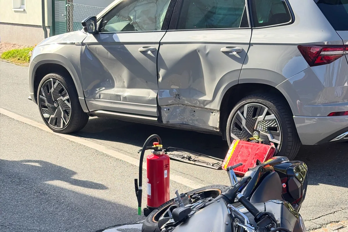 Ein silberner SUV mit Dellen an der Seite steht neben einem umgestürzten Motorrad und einem Feuerlöscher auf einer Straße.