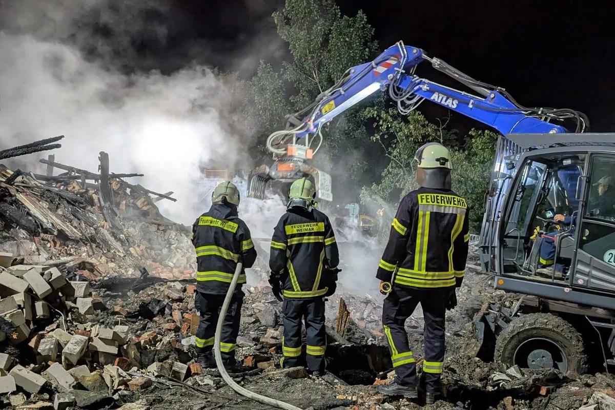 Drei Feuerwehrleute in Schutzkleidung stehen vor Trümmern, während ein Bagger bei Nacht ein Gebäude löscht.