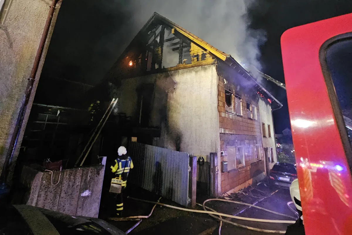 Feuerwehrleute löschen den Brand im Obergeschoss eines Hauses. Rauch steigt auf; Löschschläuche sind ausgelegt.