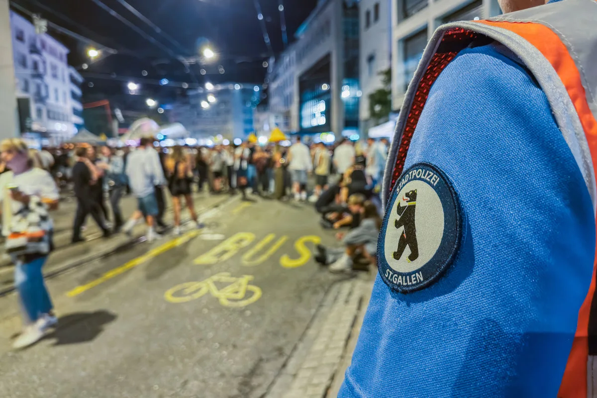 Ein Polizeibeamter in Uniform mit dem Logo der Stadtpolizei St. Gallen steht am Straßenrand und beobachtet eine große Menschenmenge bei Nacht. Im Hintergrund sind unscharf Gebäude und Straßenmarkierungen zu sehen.