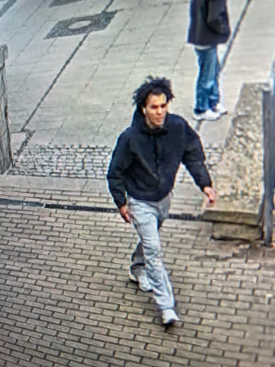 Das Bild zeigt eine Überwachungskameraaufnahme eines Mannes in dunkler Jacke und heller Hose, der auf einem gepflasterten Gehweg läuft.