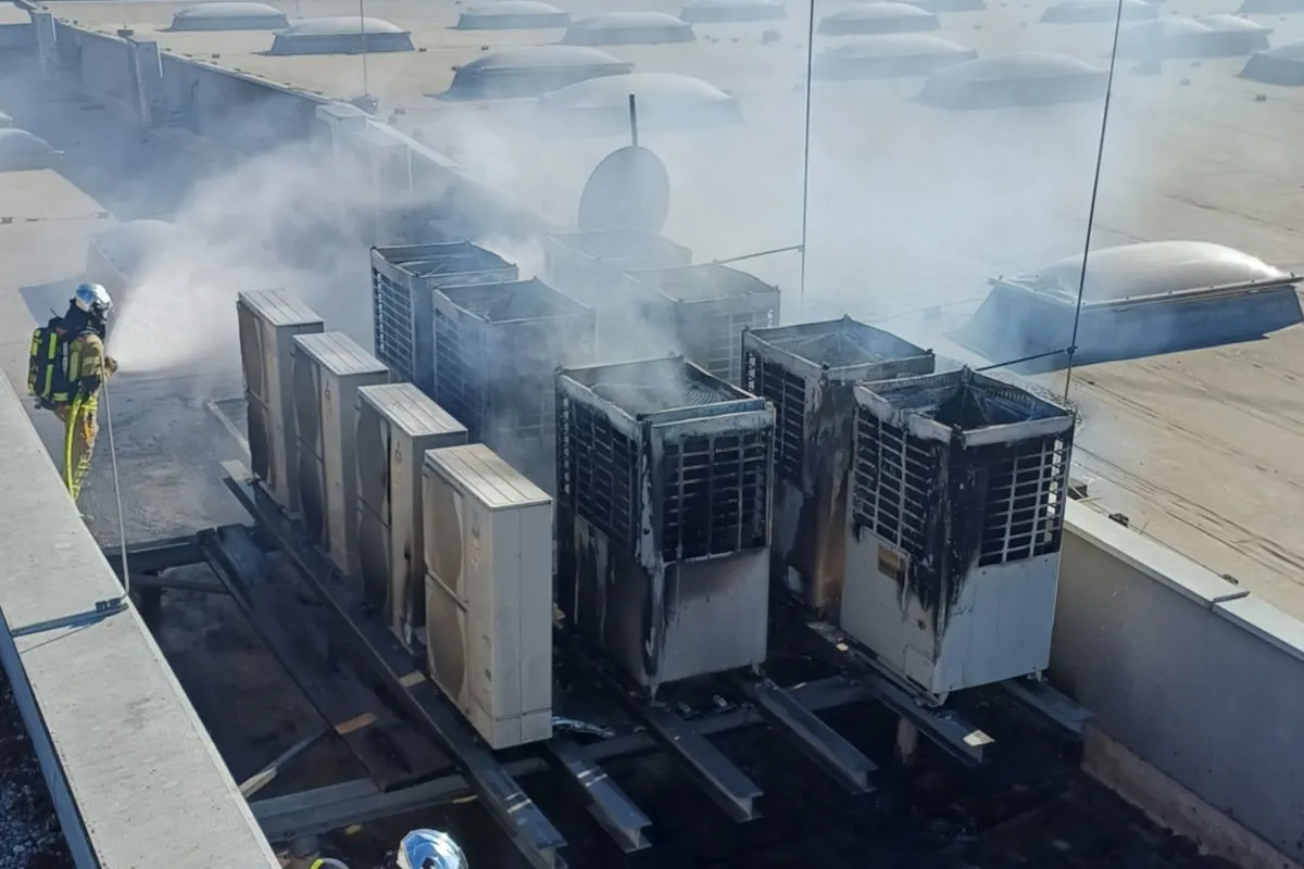 Ein Feuerwehrmann in voller Montur löscht einen Brand bei mehreren Klimaanlagen auf einem Dach. Rauch steigt aus den Geräten auf.