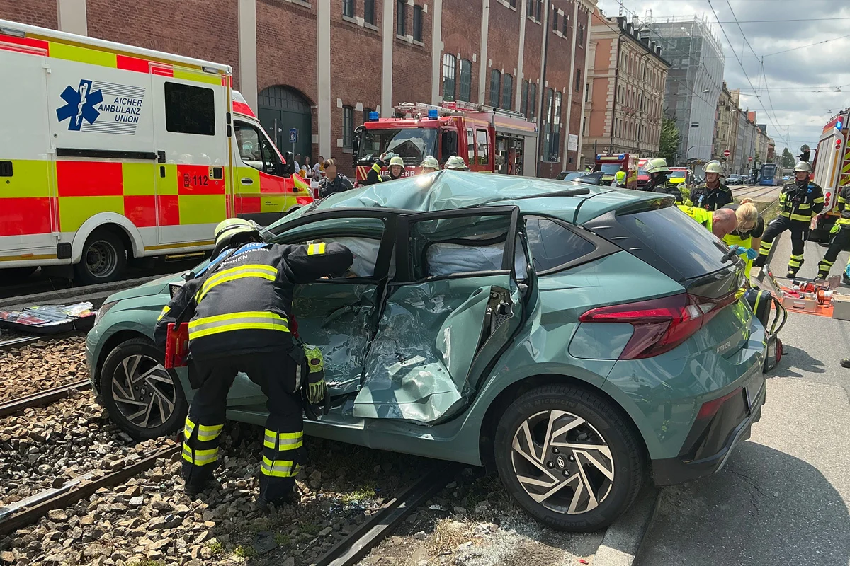 Ein Auto ist auf Tramgleisen verunglückt, umgeben von Feuerwehrleuten in Rettungsausrüstung. Ein Krankenwagen ist im Hintergrund sichtbar.