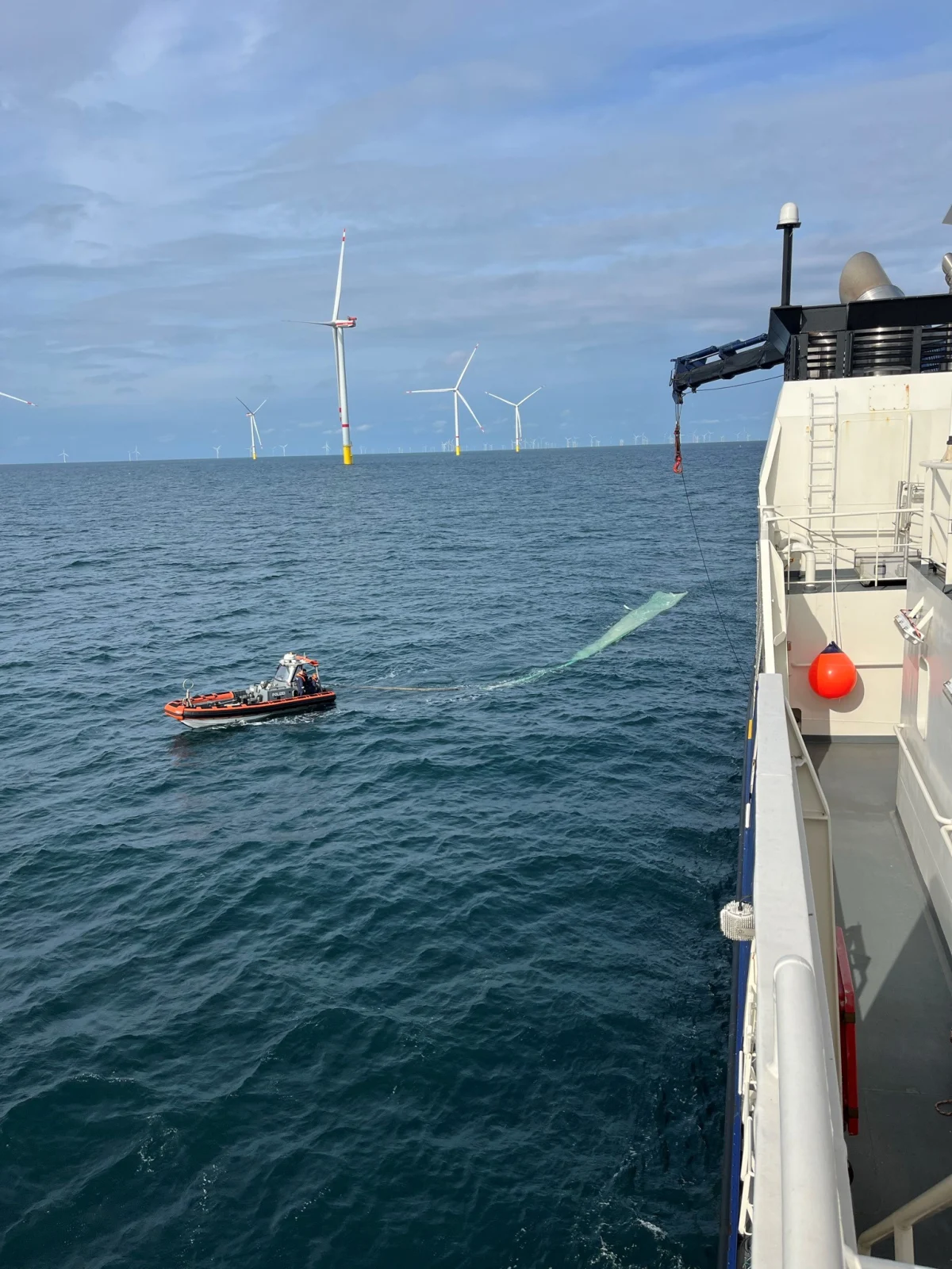 Ein Schlauchboot fährt neben einem großen Schiff auf offenem Wasser. Im Hintergrund sind mehrere Windturbinen sichtbar.