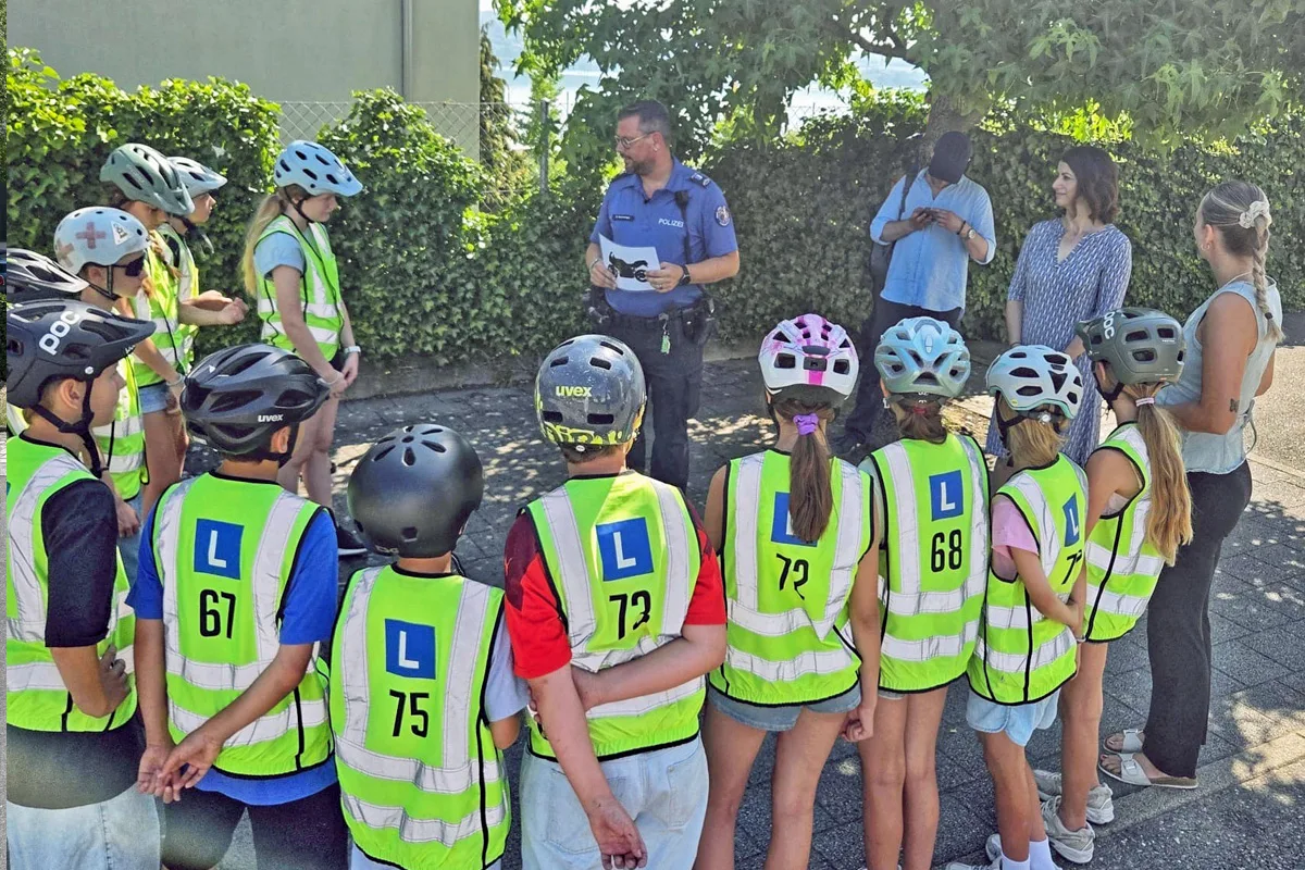 Ein Polizist steht vor einer Gruppe von Kindern, die leuchtende Westen und Helme tragen. Sie befinden sich im Freien, umgeben von Büschen.