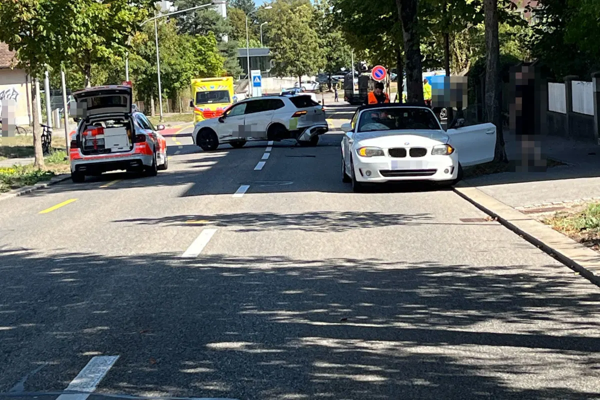 Eine Stadtstraße ist abgesperrt durch ein Polizeifahrzeug, einen Krankenwagen und ein weiteres Auto. Menschen stehen daneben.