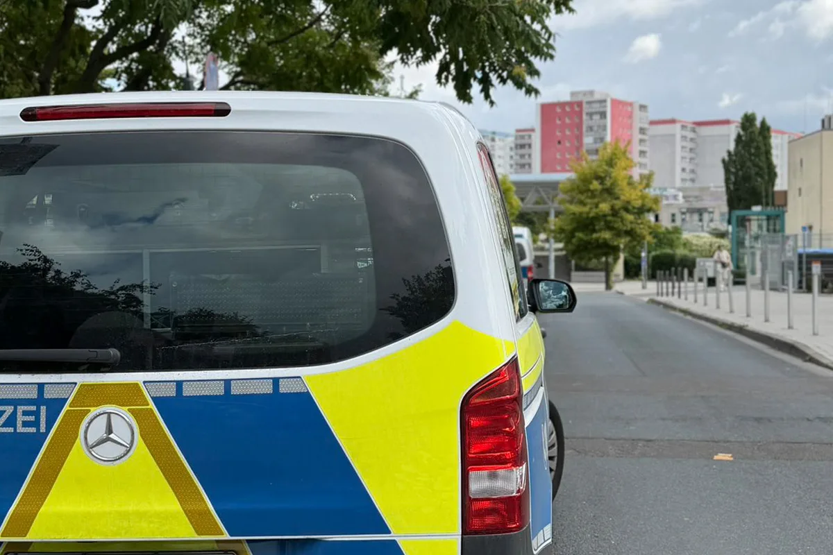 Das Bild zeigt ein Polizeifahrzeug, erkennbar an den blauen und gelben Markierungen, auf einer städtischen Straße. Im Hintergrund sind mehrstöckige Gebäude und Bäume sichtbar.