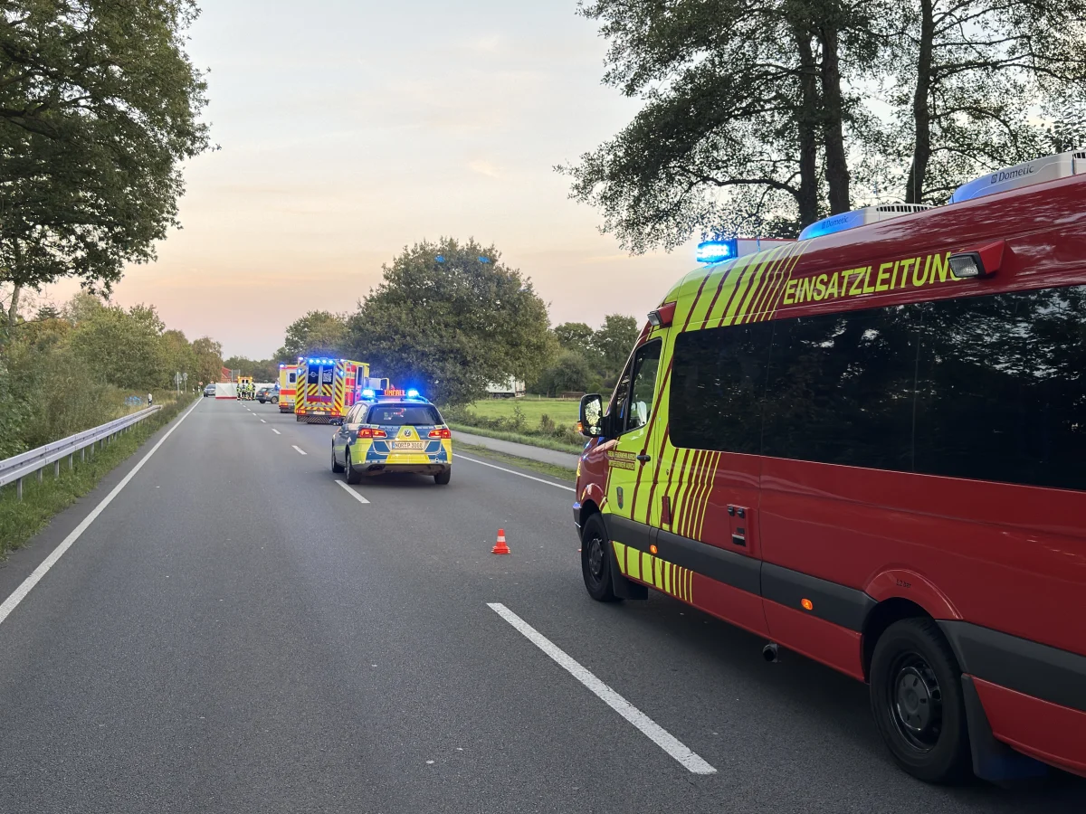 Einsatzfahrzeuge mit Blaulicht auf einer Landstraße, darunter ein rotes Fahrzeug der Einsatzleitung. Bäume säumen die Straße.