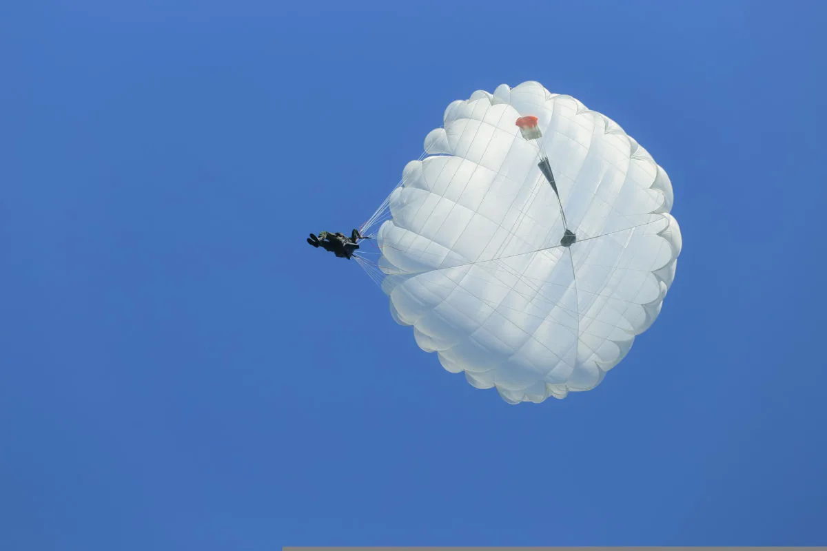 Eine Person schwebt mit einem großen, weißen Fallschirm am klaren blauen Himmel. Der Fallschirmspringer ist in der Luft deutlich sichtbar.
