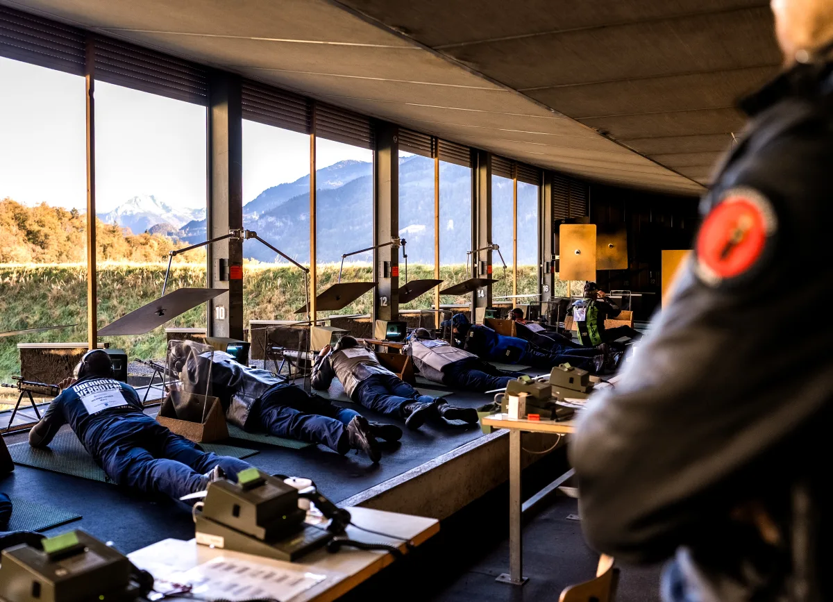 Mehrere Personen in Liegeposition auf einem Schießstand, während im Hintergrund Berge und eine Lichtung zu sehen sind.