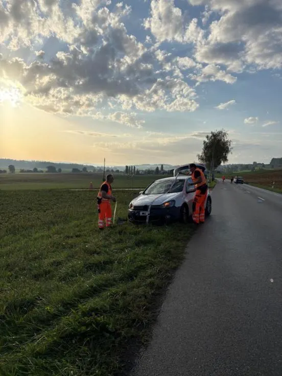 Ein Auto steht abseits einer Landstraße auf einem Feld, daneben zwei Personen in orangefarbener Kleidung.