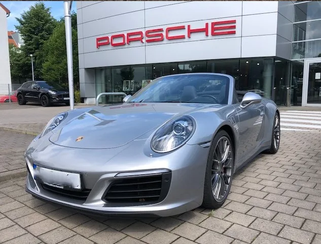 Ein silberner Porsche steht vor einem modernen Autohaus mit großen Glasfronten unter blauem Himmel.