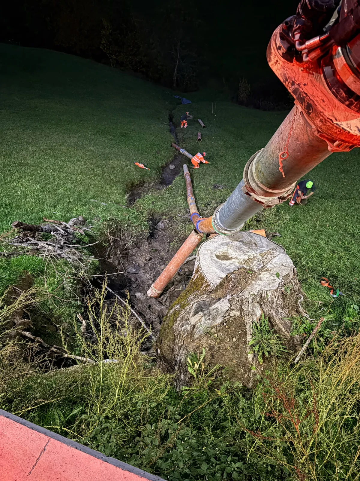 Ein dicker Schlauch wird bei Nacht über eine grüne Wiese verlegt. Mehrere Personen in orangenen Overalls arbeiten daran.