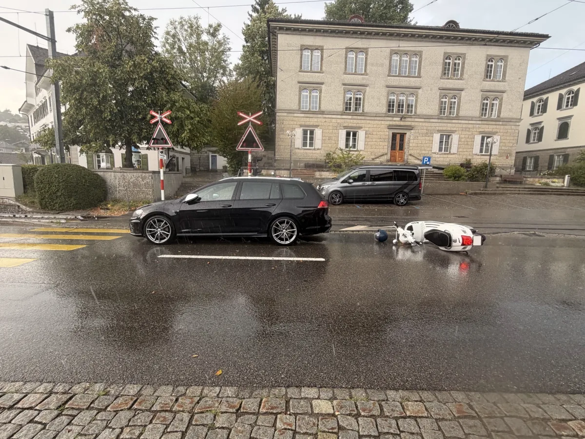 Auf einer regennassen Straße liegt ein weißes Motorrad, umgeben von einem schwarzen Auto und einem Verkehrsübergang mit Bahnzeichen im Hintergrund.