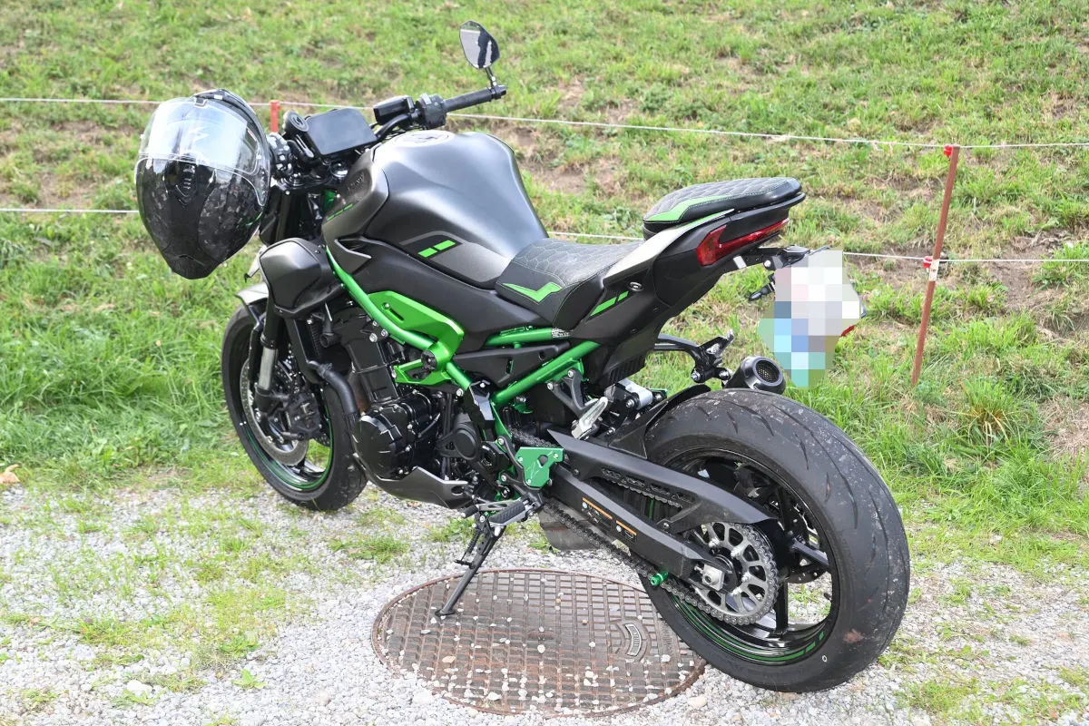 Ein schwarzes Motorrad mit grünem Rahmen steht auf einem Gitterdeckel auf einem mit Gras bewachsenen Feldweg. Der Hintergrund zeigt einen abgesperrten Wiesenbereich.