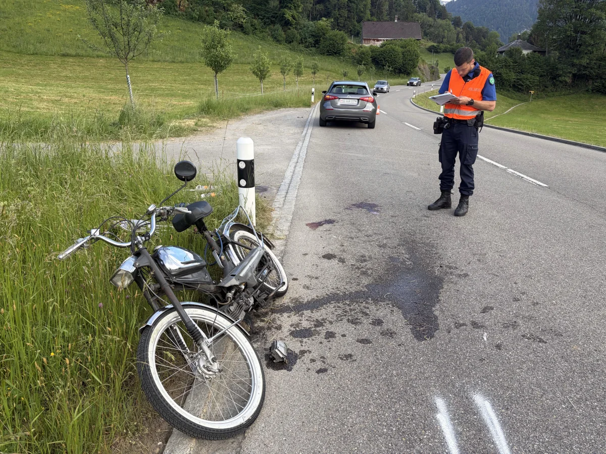 Ein beschädigtes Moped lehnt am Straßenrand an einem Pfosten. Ein Polizist in orangener Weste notiert sich Details des Unfalls auf der Straße.