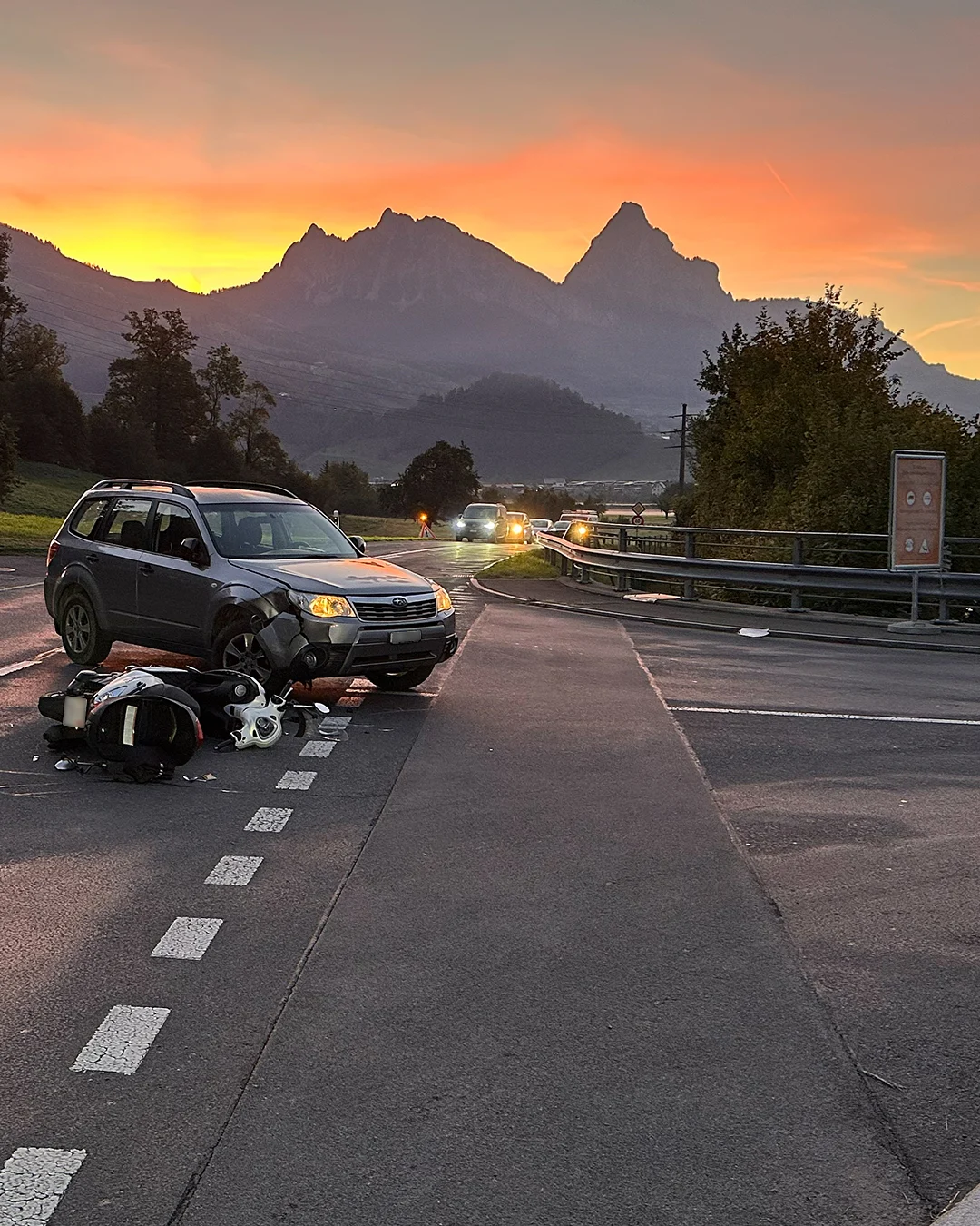 Ein silbernes Auto und ein beschädigtes Motorrad liegen auf einer Straße bei Sonnenuntergang. Im Hintergrund sind Alpen sichtbar.