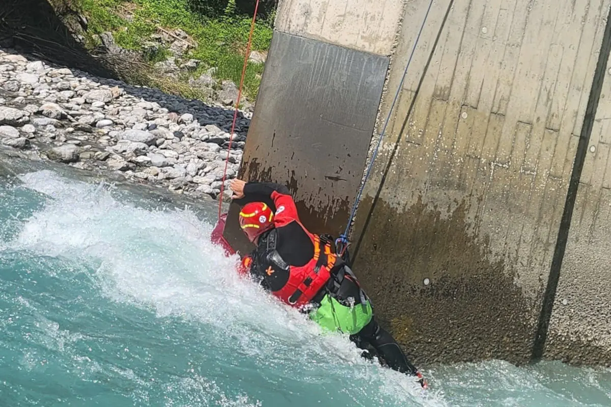 Ein Rettungsschwimmer in spezieller Ausrüstung ist an einem Seil befestigt und arbeitet in einer starken Strömung neben einem Betonpfeiler. Im Hintergrund sind Flusssteine und Vegetation zu sehen.