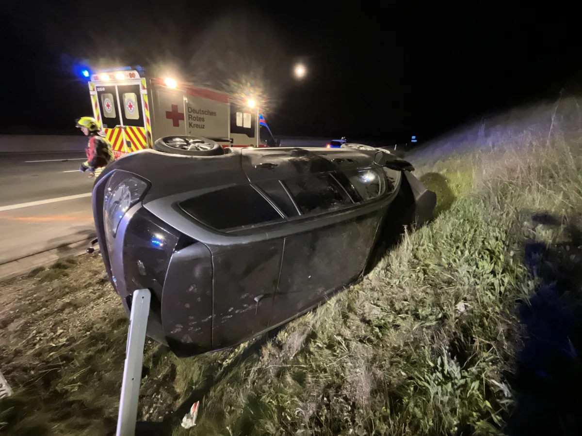Ein Auto liegt auf der Seite im Gras neben einer Autobahn. Ein Rettungsfahrzeug und ein Helfer sind sichtbar.