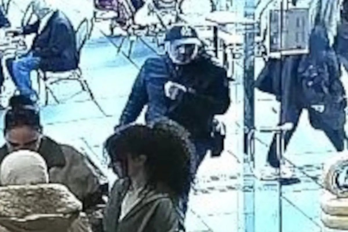 Ein Mann in dunkler Kleidung und Mütze steht in einem belebten Café, er scheint von einer Überwachungskamera erfasst worden zu sein.