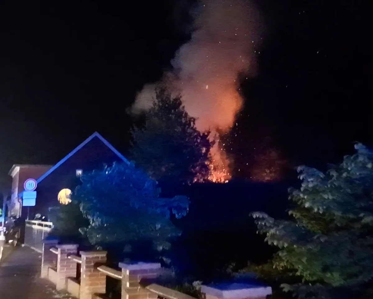 Ein Feuer brennt in der Nacht hinter einem Haus, erzeugt Rauch und leuchtet orange. Vor dem Haus sind Bäume und Zäune sichtbar.