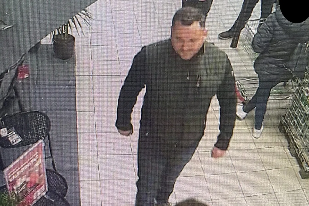 Ein Mann in dunkler Kleidung geht durch einen Einkaufsbereich mit Fliesenboden. Im Hintergrund sind weitere Personen und Einkaufswagen zu sehen.