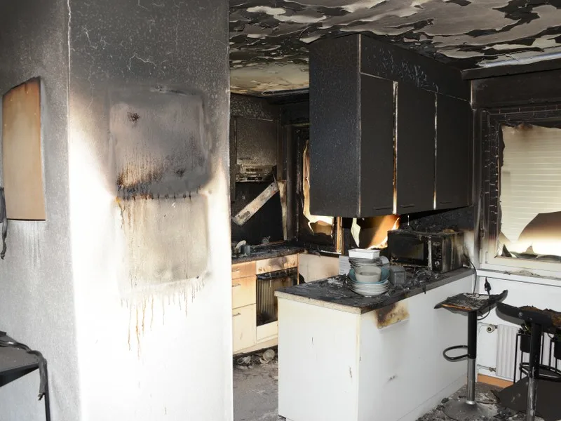 Eine stark beschädigte Küche mit verbrannten Oberflächen und Möbeln nach einem Feuer. Ruß und Asche sind sichtbar.