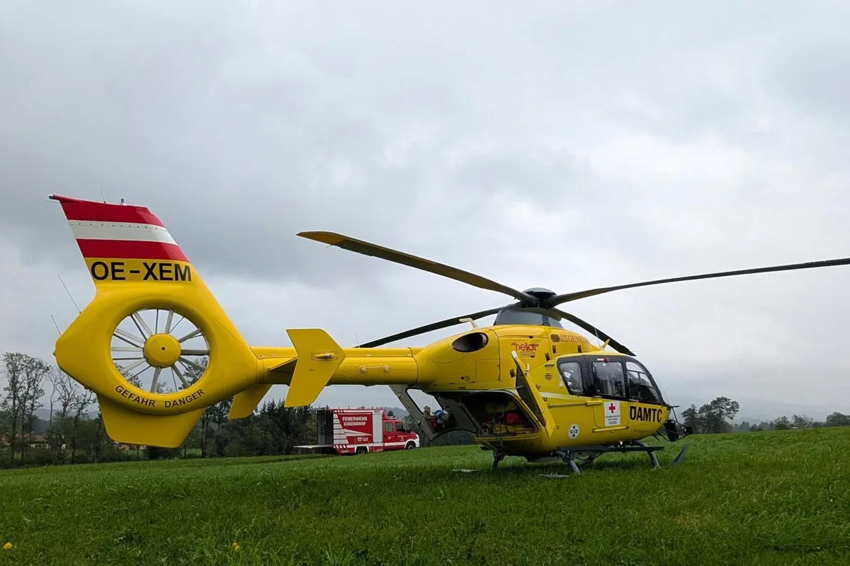 Ein gelber Rettungshubschrauber steht auf einer grünen Wiese, im Hintergrund ist ein rotes Feuerwehrfahrzeug zu sehen. Der Himmel ist bewölkt.