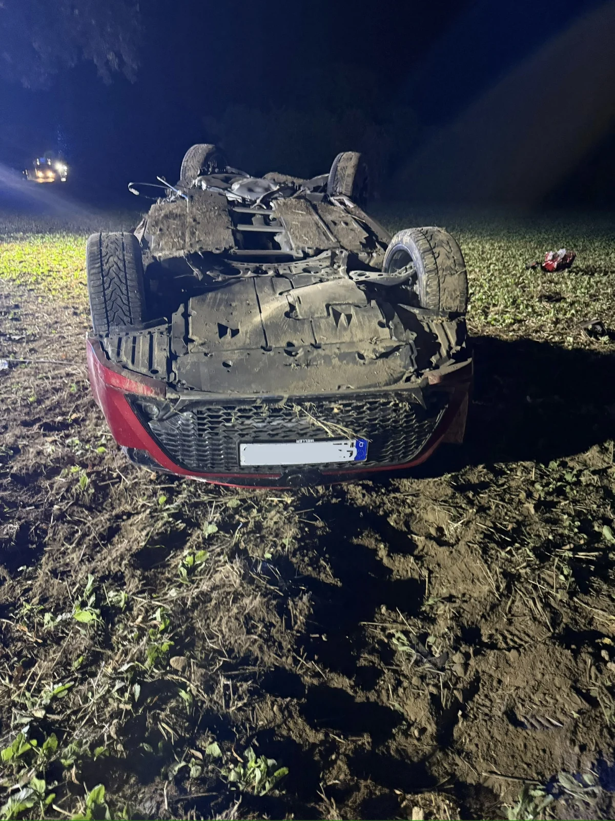 Ein rotes Auto liegt auf dem Dach auf einem Feld bei Nacht, umgeben von Erde und Pflanzen.