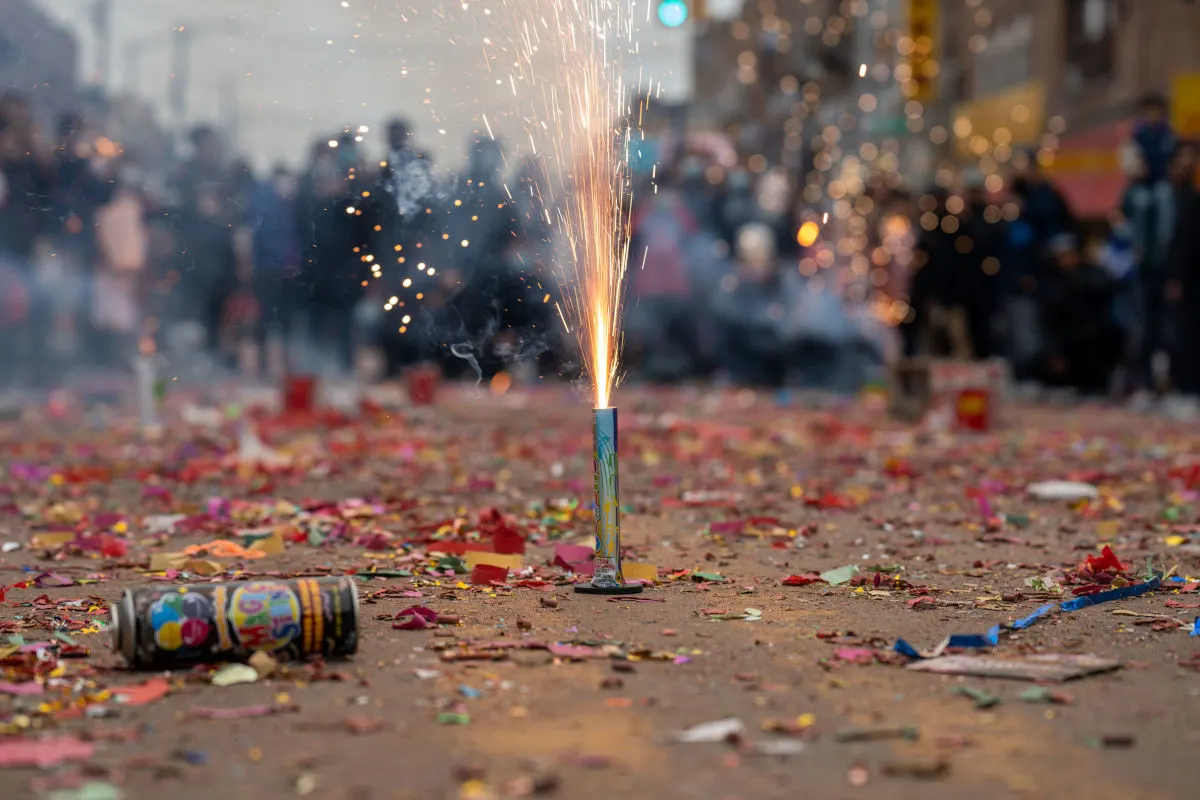 Ein Feuerwerkskörper entfaltet Funken auf einer Straße, die mit buntem Konfetti bedeckt ist. Im Hintergrund sind Menschen zu sehen.