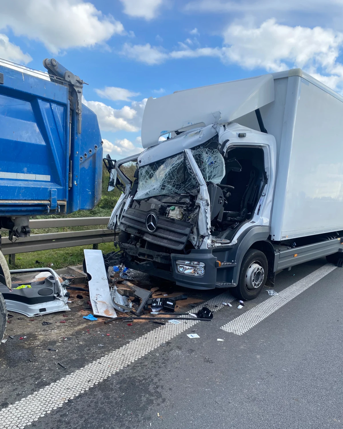 Ein weißer Lkw mit schwer beschädigter Front steht neben einem blauen Anhänger auf der Autobahn. Trümmerteile liegen verstreut auf der Straße.