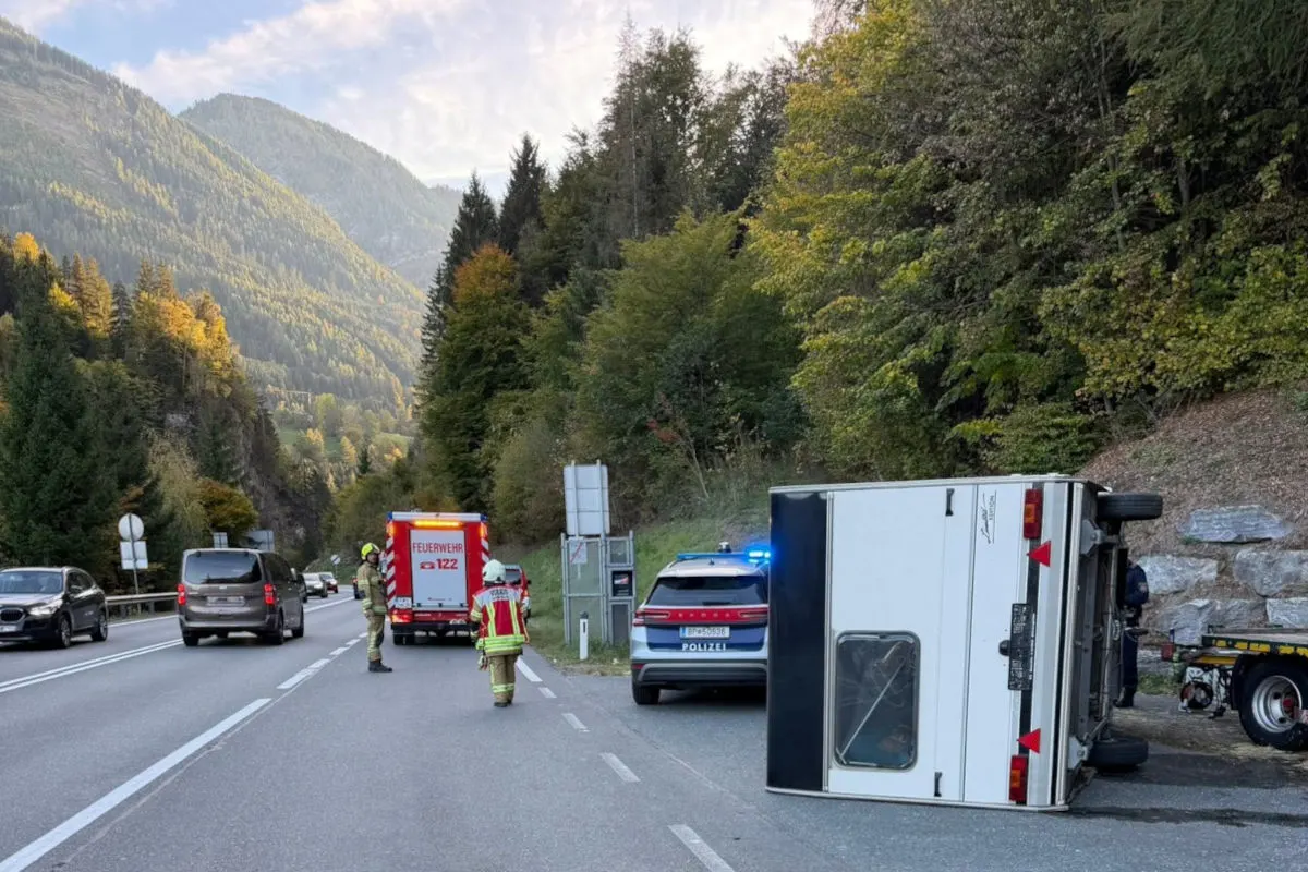 Auf dem Bild ist ein umgekippter Anhänger auf einer Landstraße zu sehen. Daneben stehen ein Polizeiwagen und ein Feuerwehrfahrzeug. Zwei Feuerwehrleute sind vor Ort.