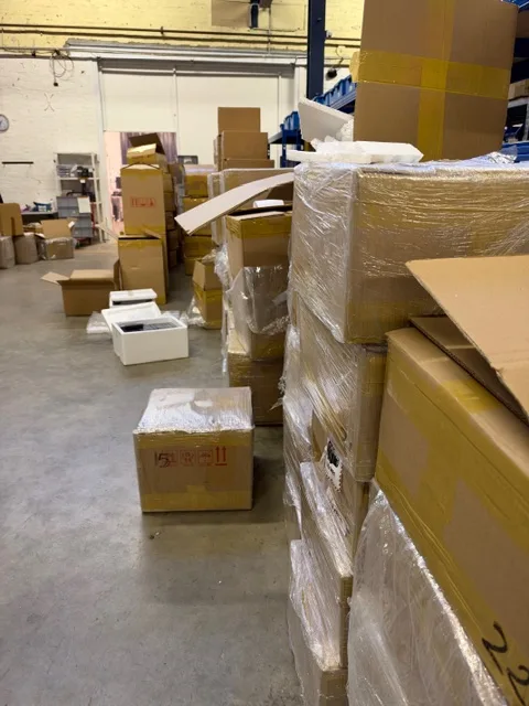 In einem Lager befinden sich zahlreiche gestapelte Kartons, einige sind offen oder ausgepackt.