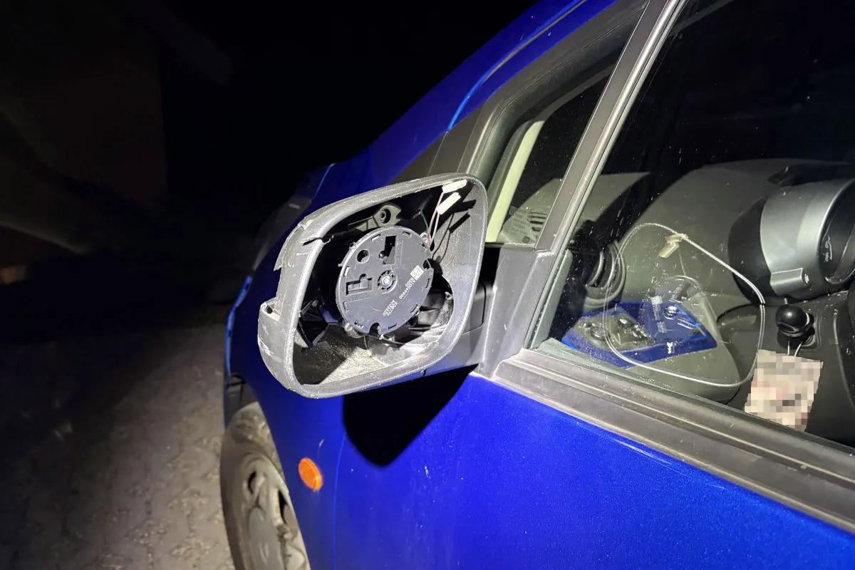 Ein blauer Wagen zeigt einen beschädigten Seitenspiegel bei Dunkelheit. Der Spiegel ist abgebrochen und die Verkabelung sichtbar.