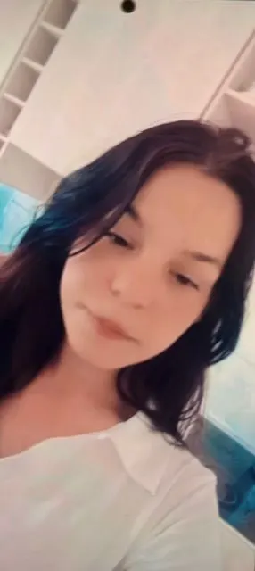 Das Bild zeigt eine Frau mit dunklen Haaren und weißem Oberteil in einem möglicherweise häuslichen Innenraum.