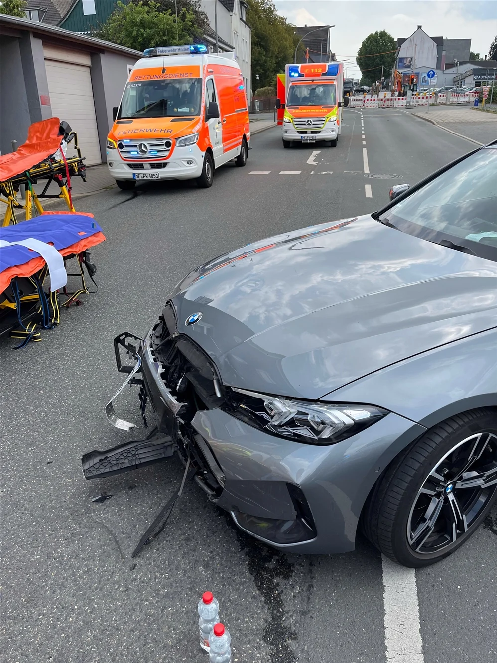 Ein beschädigter BMW steht auf einer Straße, während zwei Rettungswagen der Feuerwehr im Hintergrund sichtbar sind.