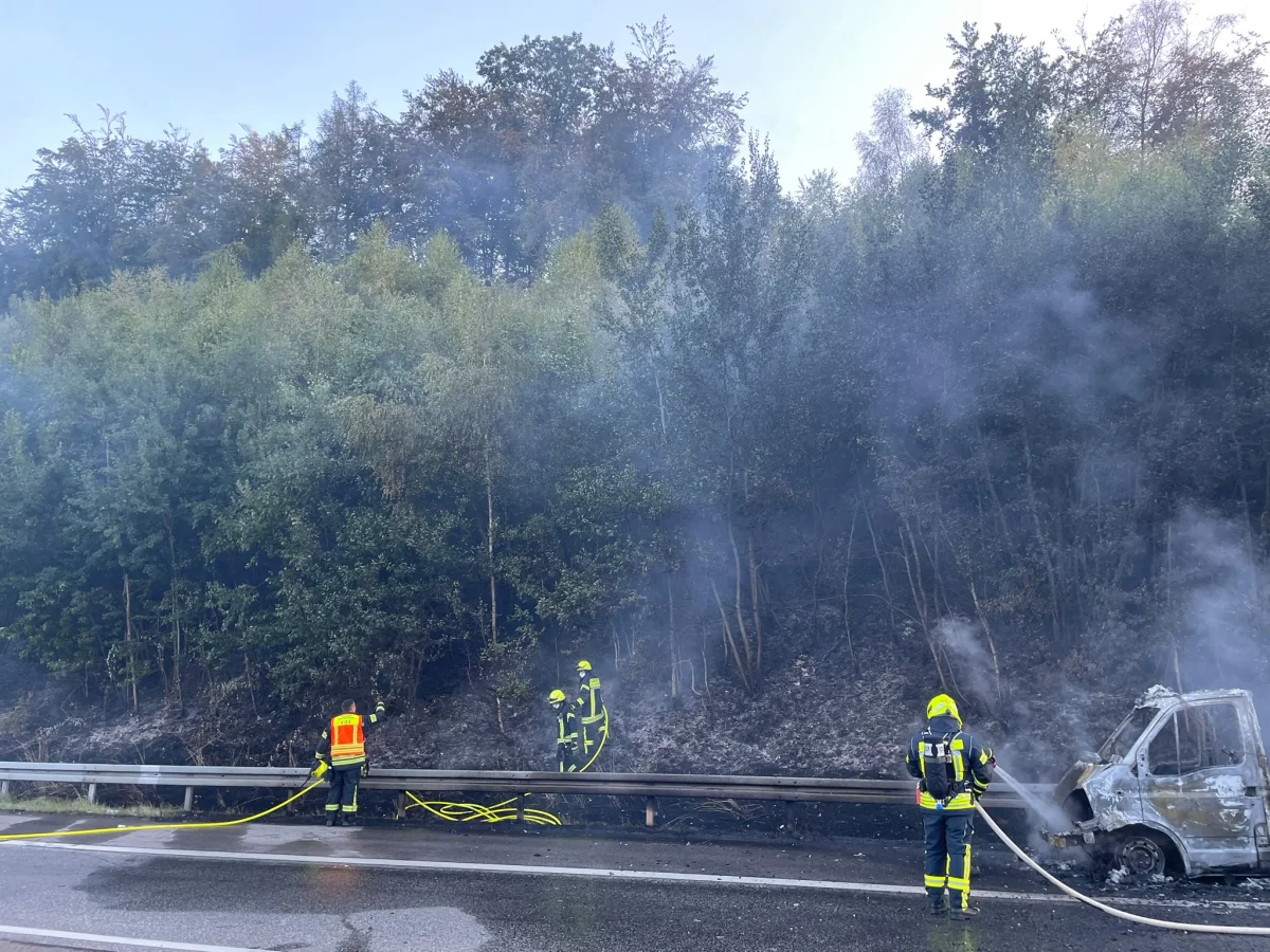 Drei Feuerwehrleute löschen ein ausgebranntes Fahrzeug auf einer Straße neben einem Waldstück. Rauch steigt auf, während die Feuerwehrschläuche Wasser spritzen.