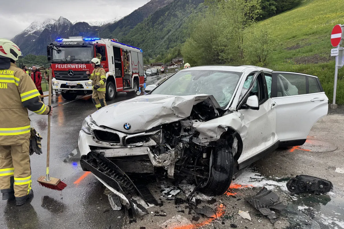 Ein stark beschädigtes weißes Auto steht auf einer nassen Straße mit geöffneter Motorhaube. Im Hintergrund ist ein Feuerwehrfahrzeug und mehrere Feuerwehrleute sichtbar.