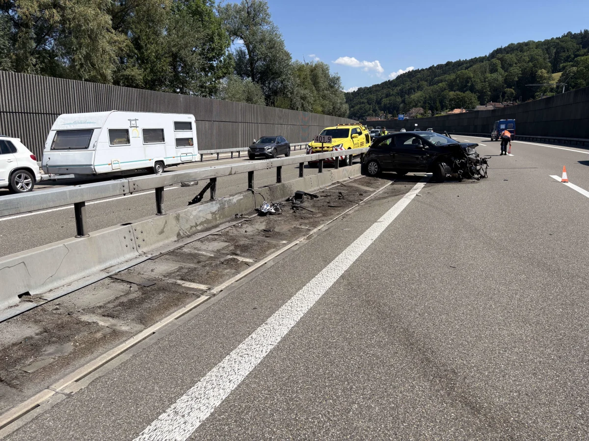 Ein schwarzes Auto steht beschädigt auf der Autobahn neben einer verbogenen Leitplanke. Einsatzfahrzeuge sind im Hintergrund sichtbar.