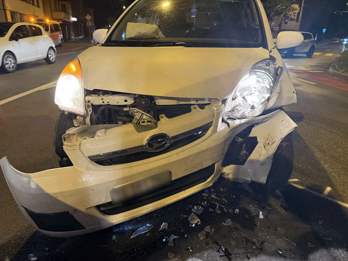 Ein weißes Auto mit stark beschädigter Frontpartie nach einem Unfall steht bei Nacht an einer Kreuzung. Glassplitter sind auf dem Boden zu sehen.