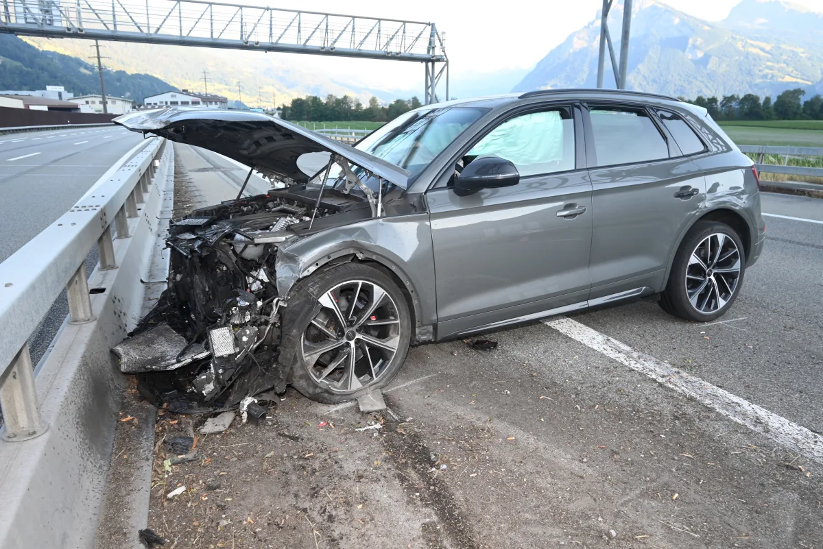 Ein graues Auto steht mit erheblichem Frontschaden an einer Leitplanke auf einer Straße. Der Motorraum ist offen und Teile liegen verstreut.